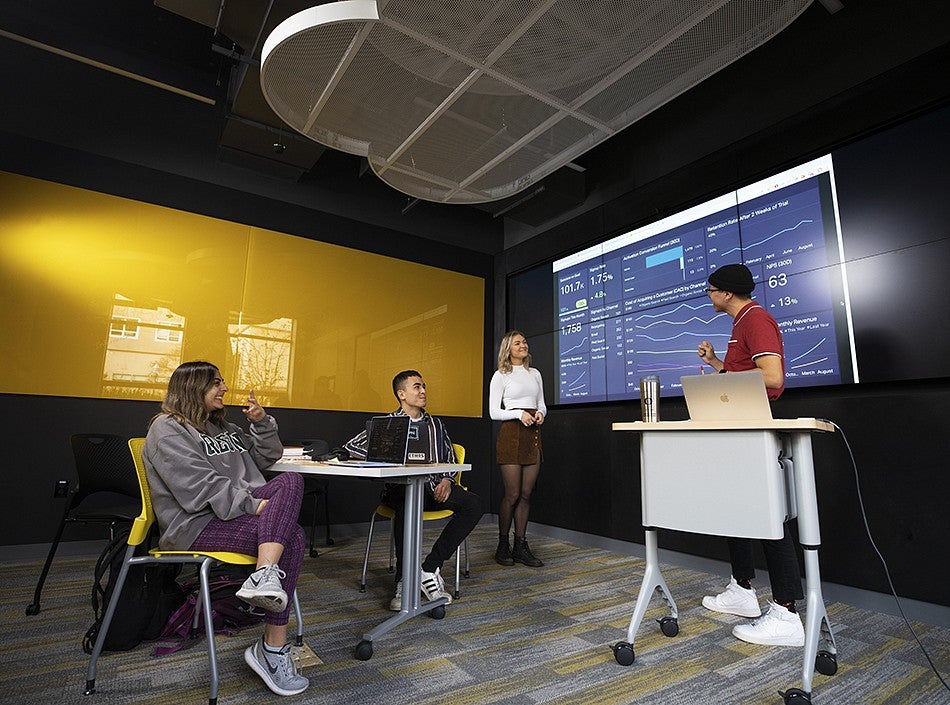Four students collaborate in a group discussion in the newly renovated social media lab. Wall of screens behind the students show graphs and other measured analytics.