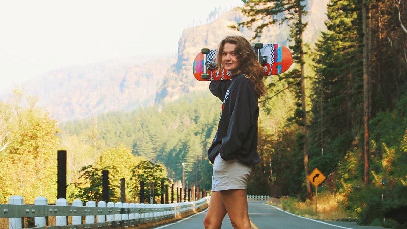 Caitlin Crowley crosses a rural highway carrying a skateboard over her shoulder