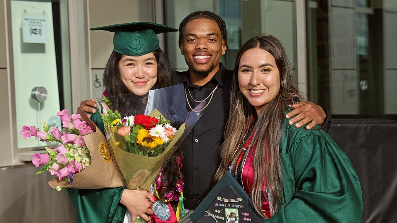 two SOJC graduates wearing green regalia pose with a third person outside of Matthew Knight Arena