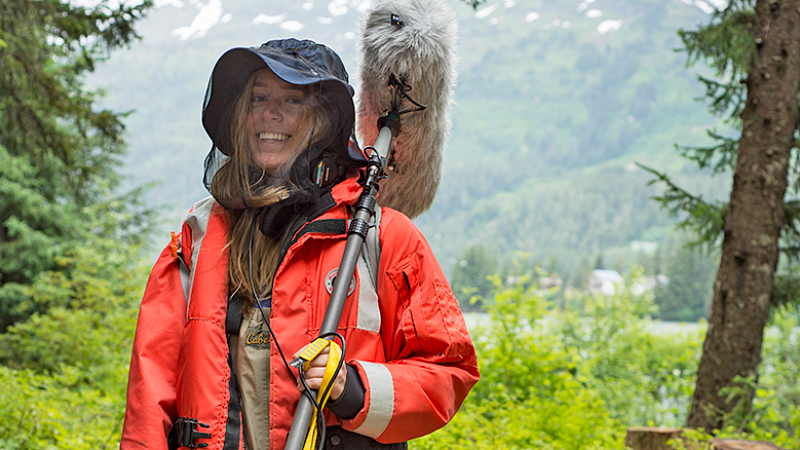 a student wearing outdoor gear holds a large boom microphone, with a wooded mountain scene in the background
