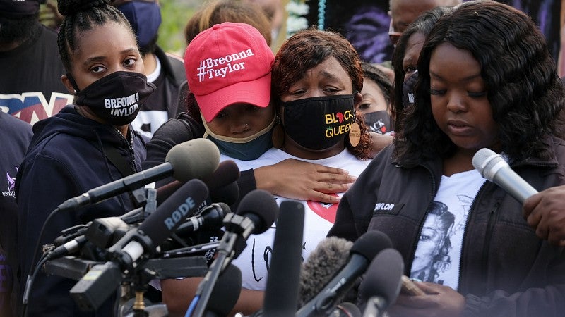 People gather around a microphone at a press conference wearing shirts reading "Breonna Taylor."