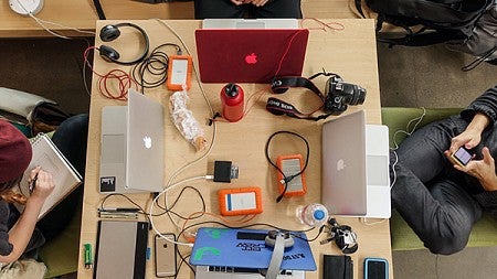 An overhead photo of a table covered in equipment including computers, cameras, headphones, and cellphones.