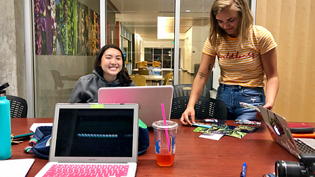 two students work at a table with laptops, a camera, and photos