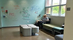 A student using a laptop studies in front of a wall covered in handwritten announcements in Allen Hall at the University of Oregon School of Journalism and Communication