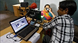 A man at a computer holds a device to the eyes of a woman. The computer screen shows a scan of her eyes.