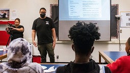 Bruce Poinsette stands in front of a classroom of students with a projector screen behind him