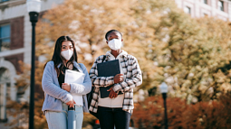 Two college students wearing masks stand together on campus (By Charlotte May from Pexels).