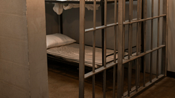 Behind prison bars lies an empty prison bed with white sheets and a single pillow (By RODNAE Productions from Pexels).