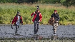 Three young men in waders walk through a stream. The middle one is carrying a camera.