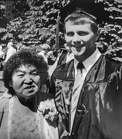 a black and white photo of Craig Harris, wearing a graduation cap and gown, standing next to his mother