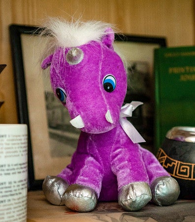 a stuffed purple unicorn children's toy sits on a bookshelf