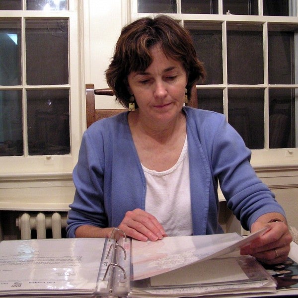 Faris reads Myra Genn's scrapbook while sitting at a table and wearing a lavender blouse. 